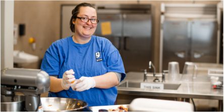 A person wearing a blue shirt and gloves prepares food in a kitchen setting
