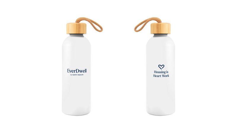 A photo of a water bottle with a bamboo cap and a strap. The Everdwell logo is on one side, and "Housing is Heart Work" is on the other.